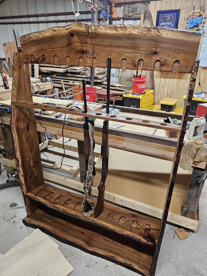 K&ouml;nnte beinhalten: Ein rustikaler Gewehrst&auml;nder aus Holz mit nat&uuml;rlicher Kante. Der St&auml;nder, mit dunkelbraunem Finish, hat Schlitze f&uuml;r mehrere Schusswaffen, wobei zwei Gewehre ausgestellt sind. Das Design hebt die nat&uuml;rliche Maserung und Kante des Holzes hervor.