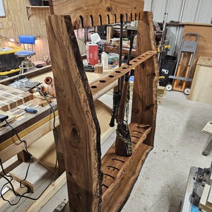 K&ouml;nnte beinhalten: Ein handgefertigter Gewehrst&auml;nder aus Holz mit einem Design mit nat&uuml;rlicher Kante. Der St&auml;nder verf&uuml;gt &uuml;ber mehrere Schlitze f&uuml;r Gewehre und ein Regal zur Aufbewahrung. Das Holz hat eine satte, dunkelbraune Farbe, die die nat&uuml;rliche Maserung und Textur zeigt. Der St&auml;nder ist f&uuml;r mehrere Schusswaffen ausgelegt.