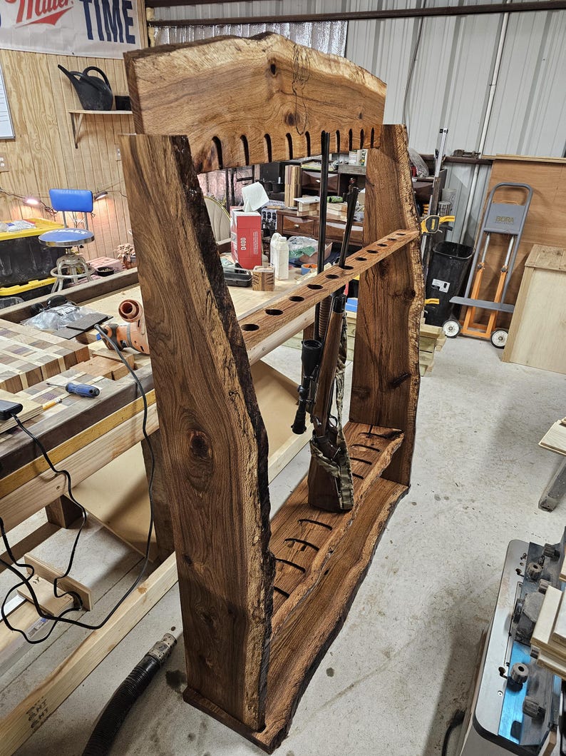 K&ouml;nnte beinhalten: Ein handgefertigter Gewehrst&auml;nder aus Holz mit einem Design mit nat&uuml;rlicher Kante. Der St&auml;nder verf&uuml;gt &uuml;ber mehrere Schlitze f&uuml;r Gewehre und ein Regal zur Aufbewahrung. Das Holz hat eine satte, dunkelbraune Farbe, die die nat&uuml;rliche Maserung und Textur zeigt. Der St&auml;nder ist f&uuml;r mehrere Schusswaffen ausgelegt.