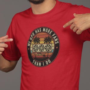 以下が含まれることがあります： ヤシの木、夕日、そして「MY GPU HAS MORE FANS THAN I DO」の文字が描かれた円形のグラフィックが特徴の赤いTシャツ。 デザインには、コンピューターファンの様式化された画像が含まれています。 シャツはブルージーンズと組み合わせています。