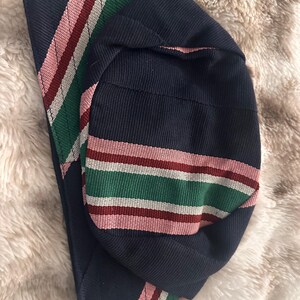 May include: A navy blue hat with a striped band in pink, green, white, and burgundy. The hat is made of a woven fabric and is folded over. The background is a soft, light-colored material.