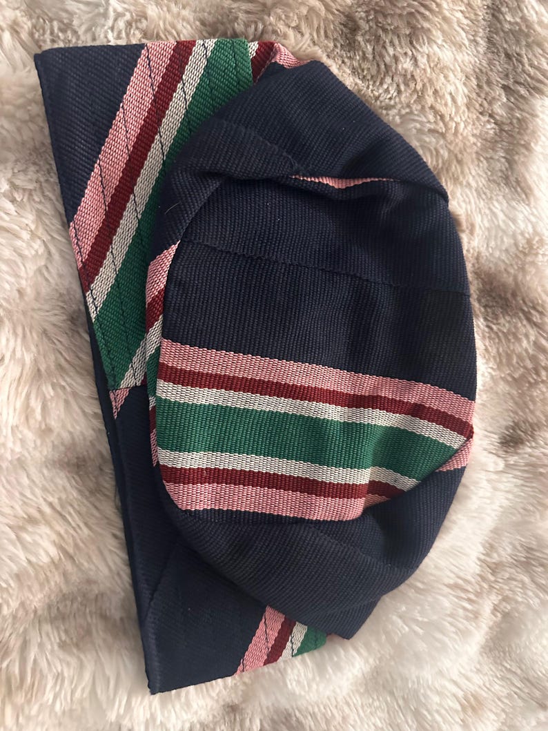 May include: A navy blue hat with a striped band in pink, green, white, and burgundy. The hat is made of a woven fabric and is folded over. The background is a soft, light-colored material.