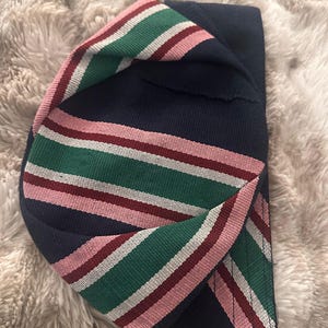 May include: A navy blue hat with a striped band in green, white, and pink. The hat is folded over, showcasing the contrasting colors and textures of the fabric. The stripes are evenly spaced.