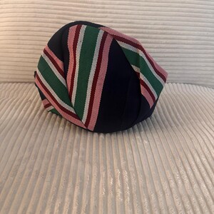 May include: A navy blue, round hat with a striped pattern in green, pink, and white. The hat is made of a soft fabric and is likely a fashion accessory. The hat is resting on a white, textured surface.