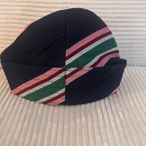May include: A navy blue hat with a striped pattern in pink, red, and green. The hat has a rounded crown and a folded brim. The stripes are diagonal across the hat. The hat is made of a textured fabric.