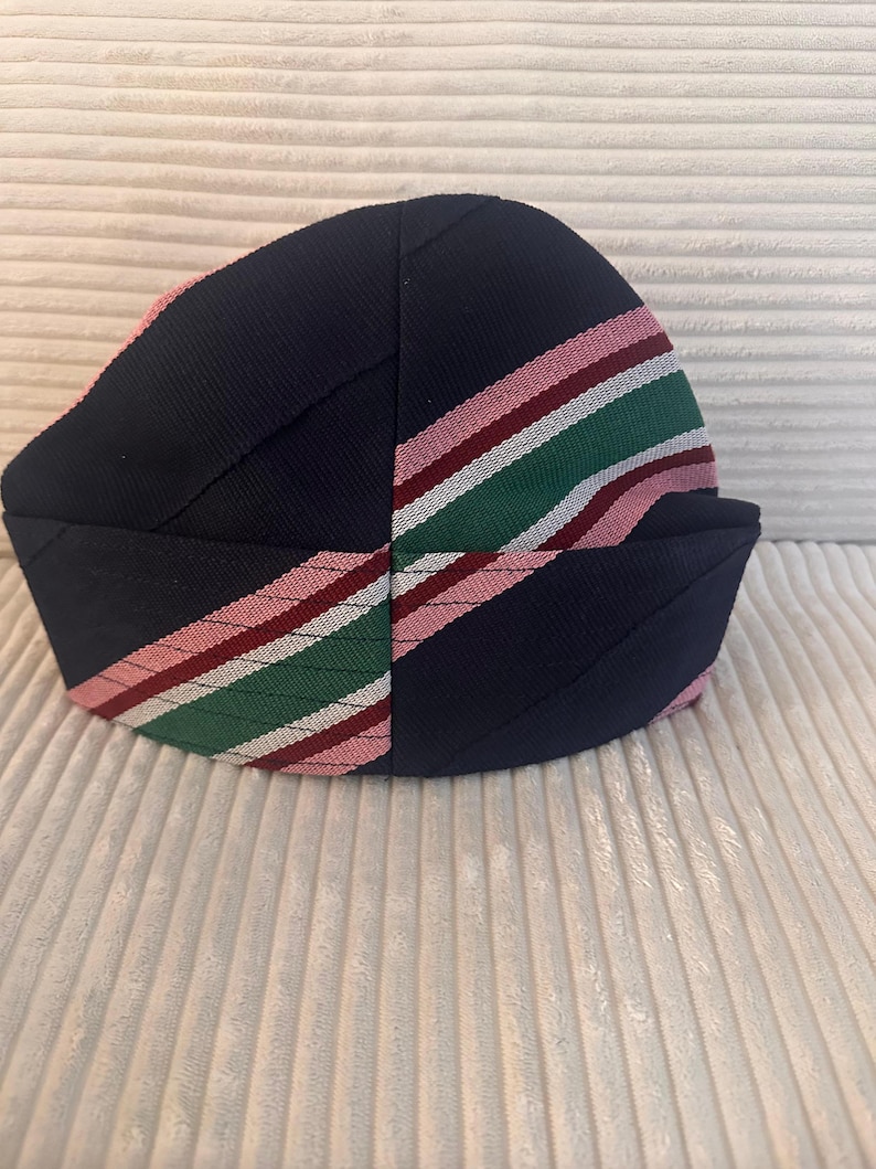 May include: A navy blue hat with a striped pattern in pink, red, and green. The hat has a rounded crown and a folded brim. The stripes are diagonal across the hat. The hat is made of a textured fabric.