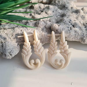 May include: A pair of off-white hoop earrings featuring a carved wheat stalk design. Each earring has a curved base with a scroll detail at the bottom and two vertical, textured wheat-like stems extending upward, secured by a thin horizontal pin.