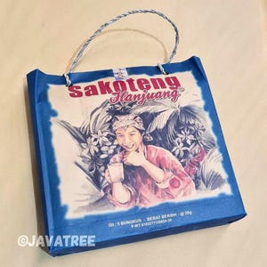 以下が含まれることがあります： 「Sakoteng Hanjuang」の文字と、グラスを持つ人物のイラストが描かれた青と白のトートバッグ。バッグにはねじれたロープの持ち手と青い縁取りがあります。イラストは熱帯の葉に囲まれています。