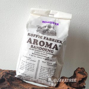 以下が含まれることがあります： 「KOFFIE FABRIEK AROMA BANDOENG」のコーヒーが入った白い紙袋。 袋には多言語のテキスト、紫色の「ROBUSTA」スタンプ、車両のグラフィックが描かれています。 袋には250グラムのコーヒーが入っています。