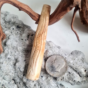 May include: A cylindrical, light tan, carved object featuring a textured, linear pattern along its length. The object is positioned next to a silver-colored United States half-dollar coin, which serves as a visual reference for its scale and size.
