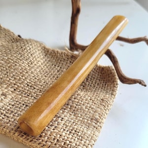May include: A cylindrical, light brown wooden object with a smooth, polished surface. The object is resting on a burlap sack, with a piece of driftwood in the background. The wood has a natural grain and a warm, inviting tone.
