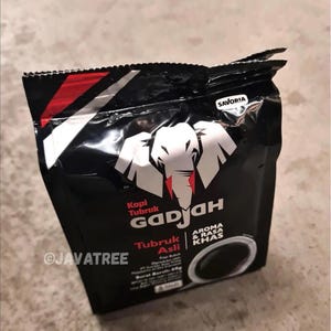 以下が含まれることがあります： 白象のロゴと赤のアクセントが付いた黒いコーヒーバッグ。「Kopi Tubruk Gadjah」と「Aroma & Rasa Khas」の文字が含まれています。バッグには「Tubruk Asli」と「SAVORIA」の文字も記載されています。