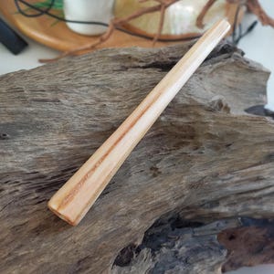 May include: A smooth, cylindrical wooden hair stick featuring a light tan color with prominent, darker brown wood grain patterns running vertically along its length. The stick has a tapered, rounded end and a polished, natural finish.