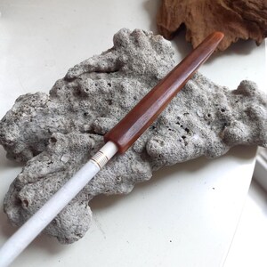 May include: A cigarette holder featuring a smooth, tapered brown wooden mouthpiece connected to a slender white stem. The junction between the brown wood and the white stem is accented with a series of thin, parallel gold-colored metallic rings.
