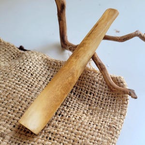 May include: A light brown, cylindrical object with a flat end, possibly a tool or decorative item. The object is resting on a burlap sack, with a piece of dried wood in the background. The item has a smooth, polished appearance.
