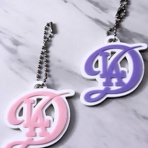 May include: Two keychains with the letters "LA" in a stylized script. One keychain is pink with a white border, and the other is purple with a white border. Each keychain has a silver ball chain attached.