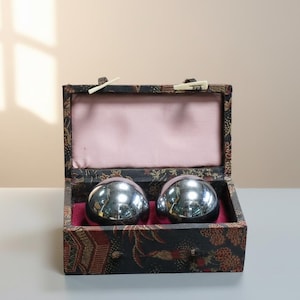 May include: A decorative box with a black and red floral pattern, containing two shiny, silver-colored metal balls. The interior of the box is lined with pink fabric, and the balls rest on a red velvet base. The box is open, revealing its contents.