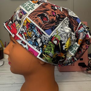 May include: A Star Wars-themed scrub cap featuring comic book panels with characters like Chewbacca, Boba Fett, and Stormtroopers. The cap has a white background with vibrant colors and the Star Wars logo.