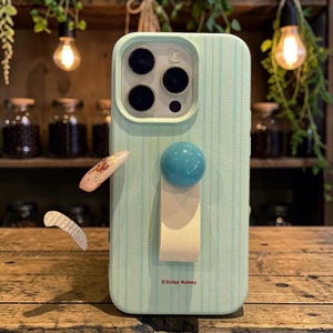 May include: A light green phone case with vertical stripes, a teal blue ball, and a cream-colored strap. The case has a camera cutout and the text "©Colss Kohey" at the bottom.