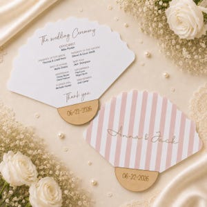 May include: Two fan-shaped wedding programs with scalloped edges. One features a white background with black text listing the ceremony details, while the other displays pink and white vertical stripes with the names Anna & Jack in script.