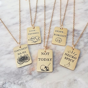 Puede incluir: Collares dorados con colgantes rectangulares. Cada colgante tiene una frase e ilustración humorística diferente, incluyendo "LOOK... SQUIRREL", "OOPS... FORGOT", "FOCUSED? HA HA", "NOT TODAY" y "NEURO SPICY".