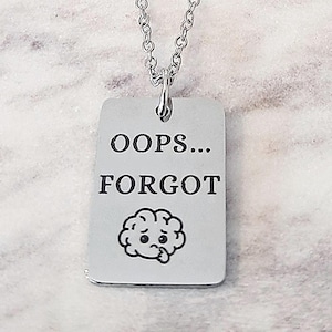 May include: A silver-colored necklace with a rectangular pendant. The pendant has the words "OOPS... FORGOT" in black text, along with a cartoon brain illustration. The chain is a delicate link style.
