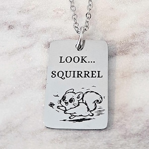 May include: A silver-colored necklace with a rectangular pendant. The pendant has the words "LOOK... SQUIRREL" printed in black, along with a cartoon squirrel illustration. The necklace chain is also silver.