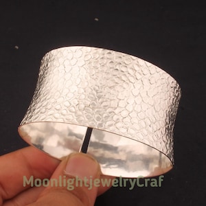 May include: A wide, silver-colored cuff bracelet with a textured, hammered surface. The bracelet has a curved shape and a split design. The text "MoonlightjewelryCraf" is visible at the bottom of the image.