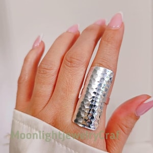 Pode incluir: Um anel grande de prata martelada com uma superfície texturizada. O anel é retangular e cobre uma parte significativa do dedo. O anel é usado em uma mão com unhas cuidadas. O texto "Moonlightjewelrycraft" é visível.