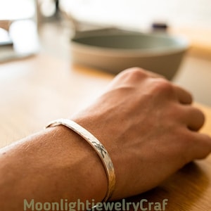 May include: A silver cuff bracelet with a hammered texture is worn on a wrist. The bracelet has a simple, elegant design. The background is blurred, showing a wooden surface and a bowl.