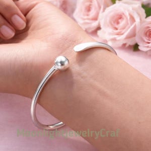 Puede incluir: Un brazalete de puño abierto de color plateado con un acento de bola. El brazalete se muestra en una muñeca, con un fondo borroso de rosas rosas. El brazalete tiene un diseño simple y elegante.