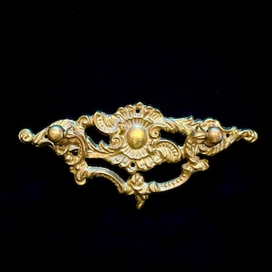 May include: Ornate gold-colored drawer pull with a symmetrical design. The hardware features a central floral motif, flanked by scrolling details and small spherical knobs on each end. The piece is set against a black background.