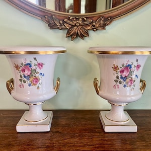 May include: Pair of white porcelain urns with gold trim and handles. Each urn features a floral design with pink, blue, and yellow flowers. The urns sit on square bases and are displayed on a wooden surface.