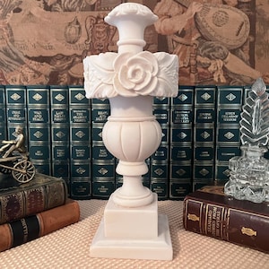 May include: A matte white alabaster urn featuring a square base, a rounded body with vertical fluting, and a decorative collar adorned with sculpted floral rose motifs and leaf patterns, topped with a flared rim and a small circular finial.