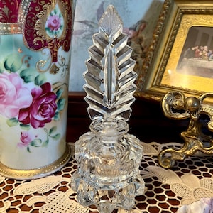 May include: A clear glass perfume bottle with a leaf-shaped stopper. The bottle has a faceted design and sits on a decorative base. The background includes a decorative vase with floral designs and a framed picture.
