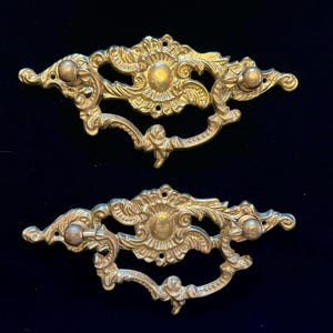 May include: Two ornate, antique brass drawer pulls. Each pull features a central floral design, curved handle, and three small knobs. The pulls have a detailed, vintage aesthetic, perfect for adding a touch of elegance to furniture.