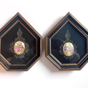 May include: Two pentagonal wall plaques featuring black frames with gold-toned inner edges. Each plaque contains a central oval porcelain medallion depicting a colorful 18th-century scene of figures, mounted on a black background with ornate filigree.