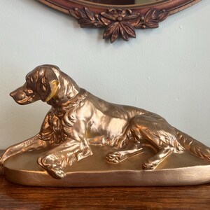 May include: A bronze-colored dog statue in a reclining pose. The dog is detailed with visible fur and facial features, resting on a matching oval base. The statue is placed on a wooden surface, with a decorative mirror in the background.
