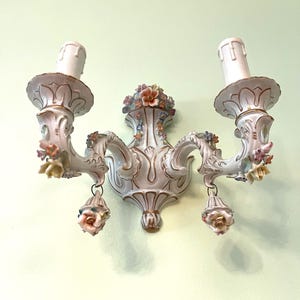 May include: A white porcelain wall sconce with two candle holders. The sconce is decorated with floral accents in pink, yellow, and blue. The sconce has a gold trim and is mounted on a light green wall. The sconce is an elegant piece of home decor.