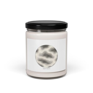 May include: A white candle jar with a black lid. The jar has a white label with a circular topographic map design in shades of grey and beige. The coordinates 34.527 N 118.247 W are printed below the map.