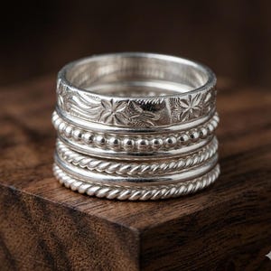 May include: A stack of silver rings with various textures. The rings include floral patterns, beaded designs, and rope-like twists. The rings are stacked on top of each other, creating a layered effect. The rings are made of a shiny, metallic material.