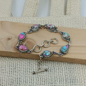 May include: A silver bracelet with teardrop-shaped turquoise, pink, and white stones. The bracelet has a toggle clasp and is displayed on a wooden surface. The text "Silver925ringshop" is visible on the bracelet.