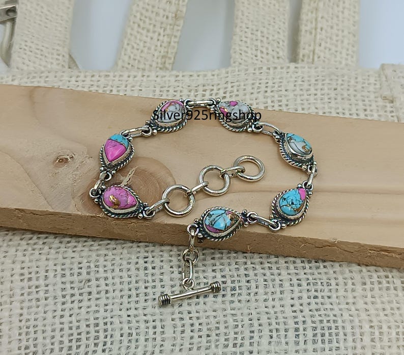 May include: A silver bracelet with teardrop-shaped turquoise, pink, and white stones. The bracelet has a toggle clasp and is displayed on a wooden surface. The text "Silver925ringshop" is visible on the bracelet.