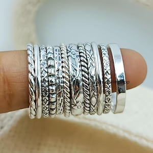Könnte beinhalten: Eine Sammlung von elf Silberringen, die zusammen an einem Finger getragen werden. Die Ringe weisen verschiedene Texturen und Muster auf, darunter gedrehte Kordeldesigns, florale Gravuren, geometrische Kreuzschraffuren und glatte Oberflächen.