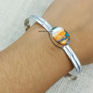 Könnte beinhalten: Ein silberner Armreif mit doppeltem Banddesign und einem zentralen ovalen Stein mit wirbelnden Mustern in Weiß, Orange und Türkisblau. Das Metall hat eine polierte Oberfläche und der Text silver925ringshop ist auf dem Band zu sehen.