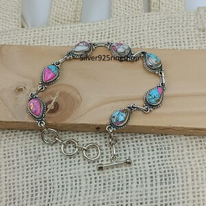 May include: A silver bracelet with teardrop-shaped stones in shades of pink, turquoise, and white. The stones are set in a silver frame with a twisted rope design. The bracelet has a toggle clasp.