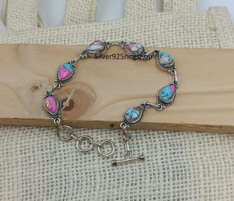May include: A silver bracelet with teardrop-shaped stones in shades of pink, turquoise, and white. The stones are set in a silver frame with a twisted rope design. The bracelet has a toggle clasp.
