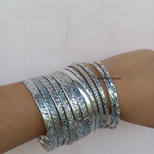 May include: A set of nine silver bangles featuring various intricate embossed patterns including floral motifs, geometric braids, and smooth polished surfaces. The bracelets are stacked together and display the text Silver926ringshop.