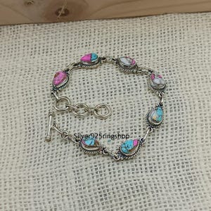 May include: A silver bracelet featuring teardrop-shaped stones in shades of pink, turquoise, and white. The stones are set in a silver frame and connected by a chain. The bracelet has a toggle clasp. The text "Silver925ringshop" is visible.
