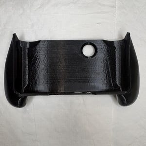 May include: A black 3D-printed controller grip accessory featuring a rectangular central housing with curved side handles. The surface displays horizontal layer lines from the printing process, and a circular cutout is positioned on the top right.